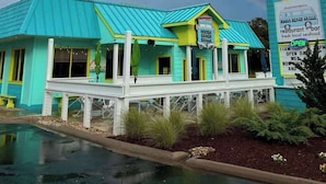 Un bâtiment turquoise avec un porche blanc, une enseigne indiquant le « Noosa Beach Grill », et un arc-en-ciel dans le ciel.