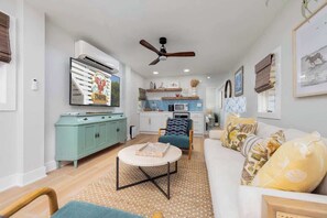 Sala moderna com sofá, mesa de centro e área de jantar com mesa e cadeiras. Cozinha ao fundo, com armários e micro-ondas.