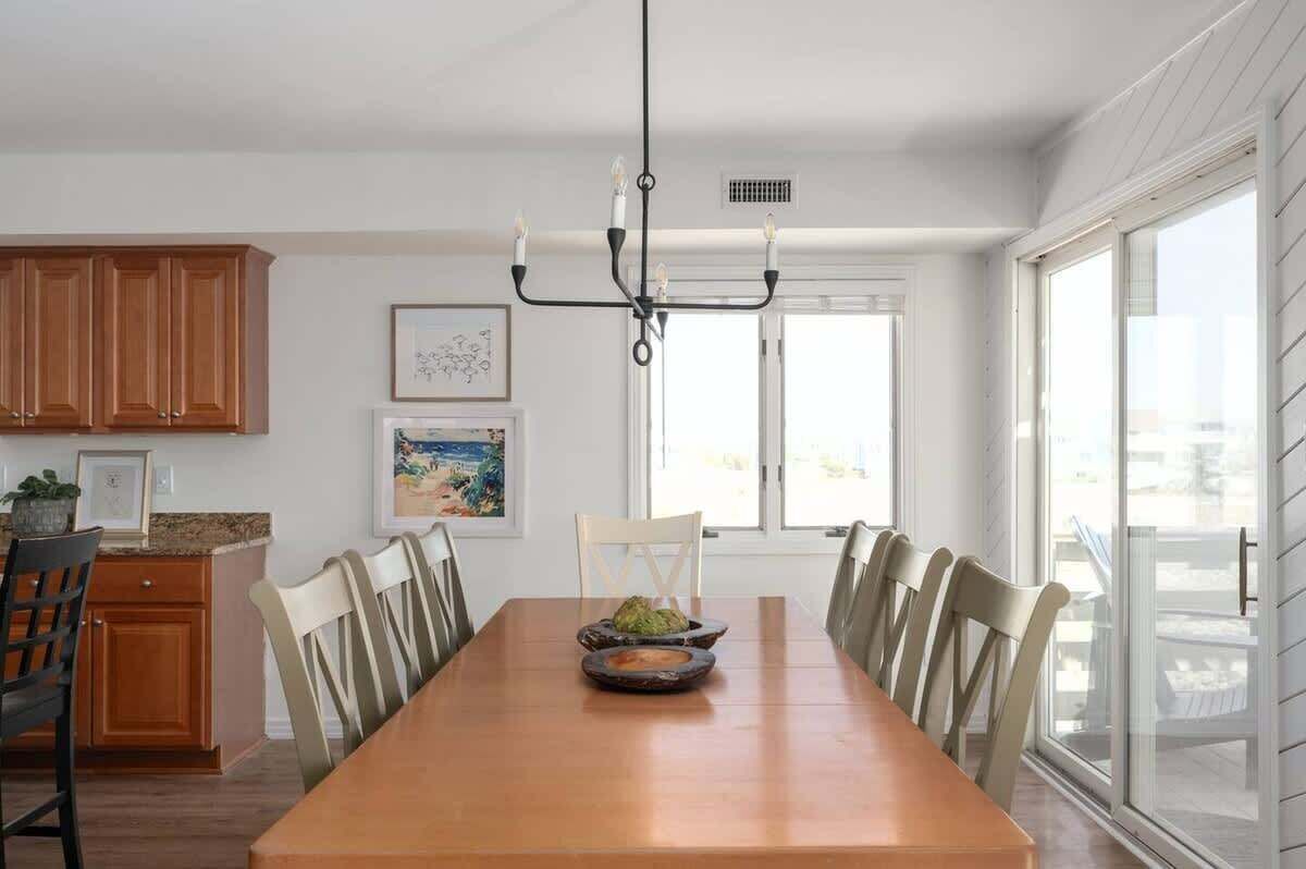A dining area with a long wooden table, six chairs, a chandelier, and large windows.
