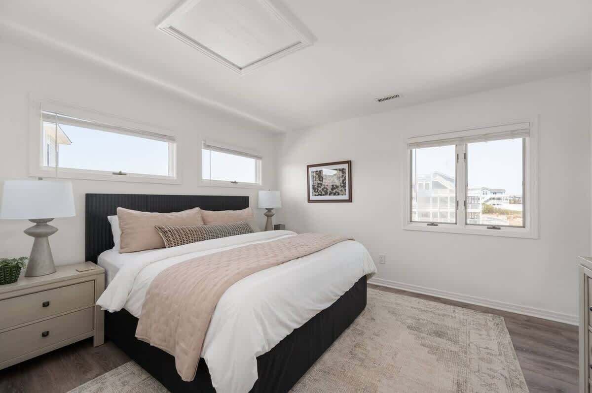 Une chambre à coucher avec un grand lit, deux tables de chevet, un luminaire au plafond et deux fenêtres donnant sur des immeubles.