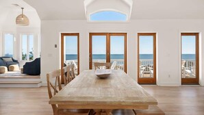 A spacious dining area with a wooden table and chairs, large windows with ocean views, and a high ceiling with a skylight. - Oceanfront, Pool & Hot Tub, Game Room, EV Charger (Corolla)