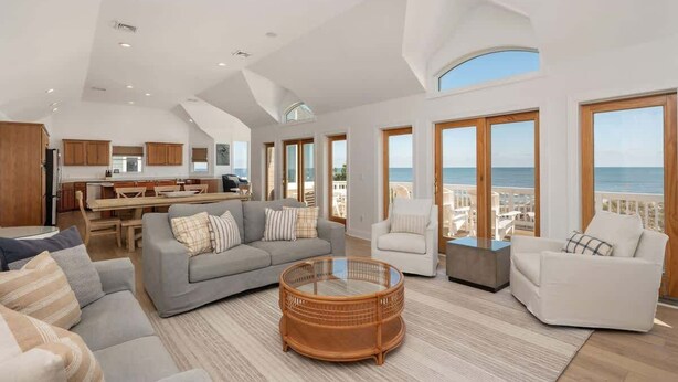 A spacious living area with a large window overlooking the sea, a dining area with wooden furniture, and a kitchen with modern appliances. - Oceanfront, Pool & Hot Tub, Game Room, EV Charger (Corolla)