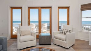 A spacious living room with a view of the ocean, featuring two white armchairs, a glass coffee table, and a large sliding glass door leading to an outdoor seating area. - Oceanfront, Pool & Hot Tub, Game Room, EV Charger (Corolla)