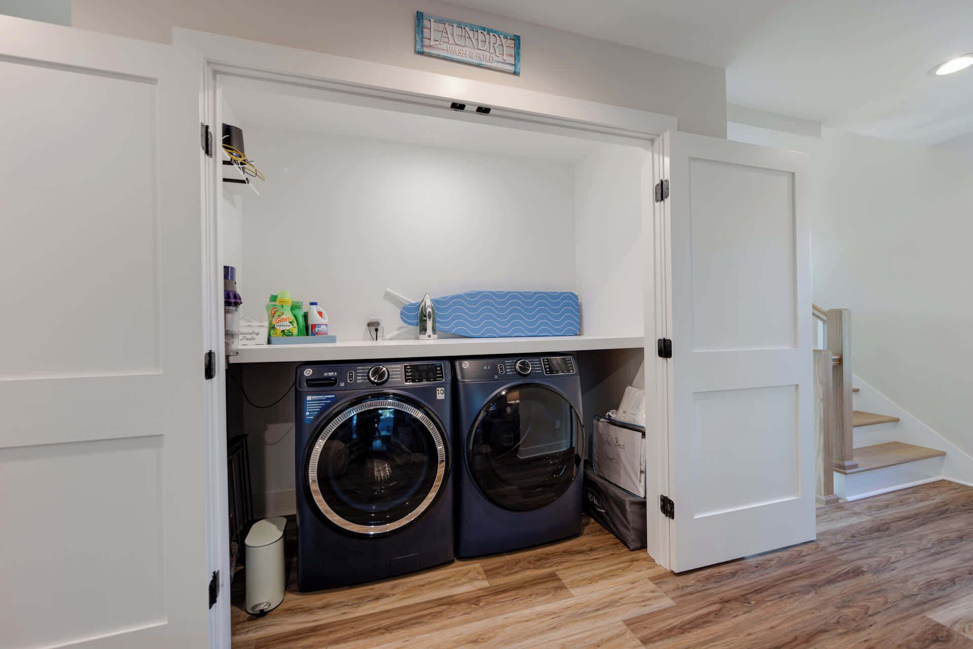 A compact laundry room with two front-loading washing machines, a drying rack, and a shelf with laundry supplies.