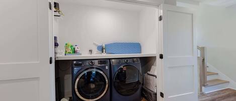 A compact laundry room with two front-loading washing machines, a drying rack, and a shelf with laundry supplies.