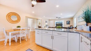 A modern kitchen with white cabinetry, a granite countertop, and a dining area with a wooden table and white chairs. - Cozy Soundside Cottage  • Bring Your Dog! (Kill Devil Hills)