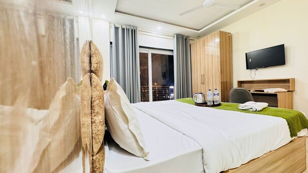 A hotel room with a bed, a flat-screen TV, a coffee maker, and a view of the city at night. - Sky Hotel (Gurugram)