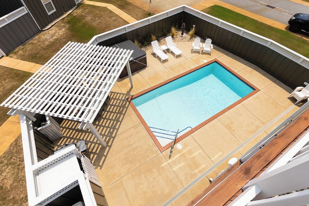A rectangular swimming pool with a white deck and lounge chairs, surrounded by a wooden fence and a pergola.