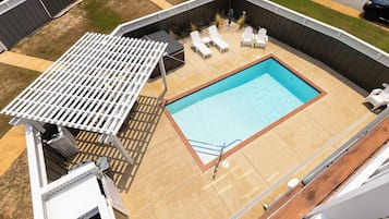 A rectangular swimming pool with a white deck and lounge chairs, surrounded by a wooden fence and a pergola.
