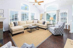 A living room with a sectional sofa, a coffee table, a patterned armchair, and a ceiling fan. - Dog Friendly Gorgeous Corolla Home Pool & Hot Tub (Corolla)