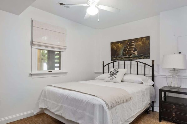 A bedroom with a bed, a ceiling fan, a window with blinds, a painting on the wall, and a bedside table with a lamp.