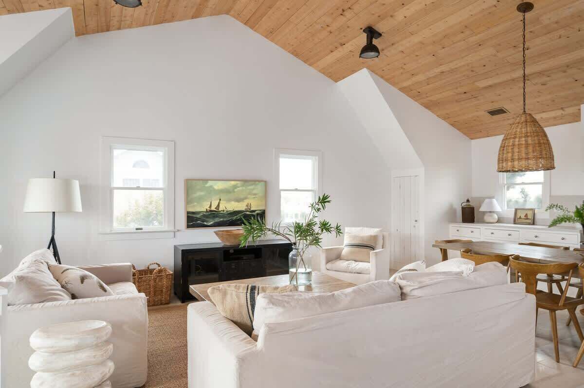 A spacious living room with a high ceiling, white walls, and a large window. There is a television, a dining table with chairs, and a sofa.