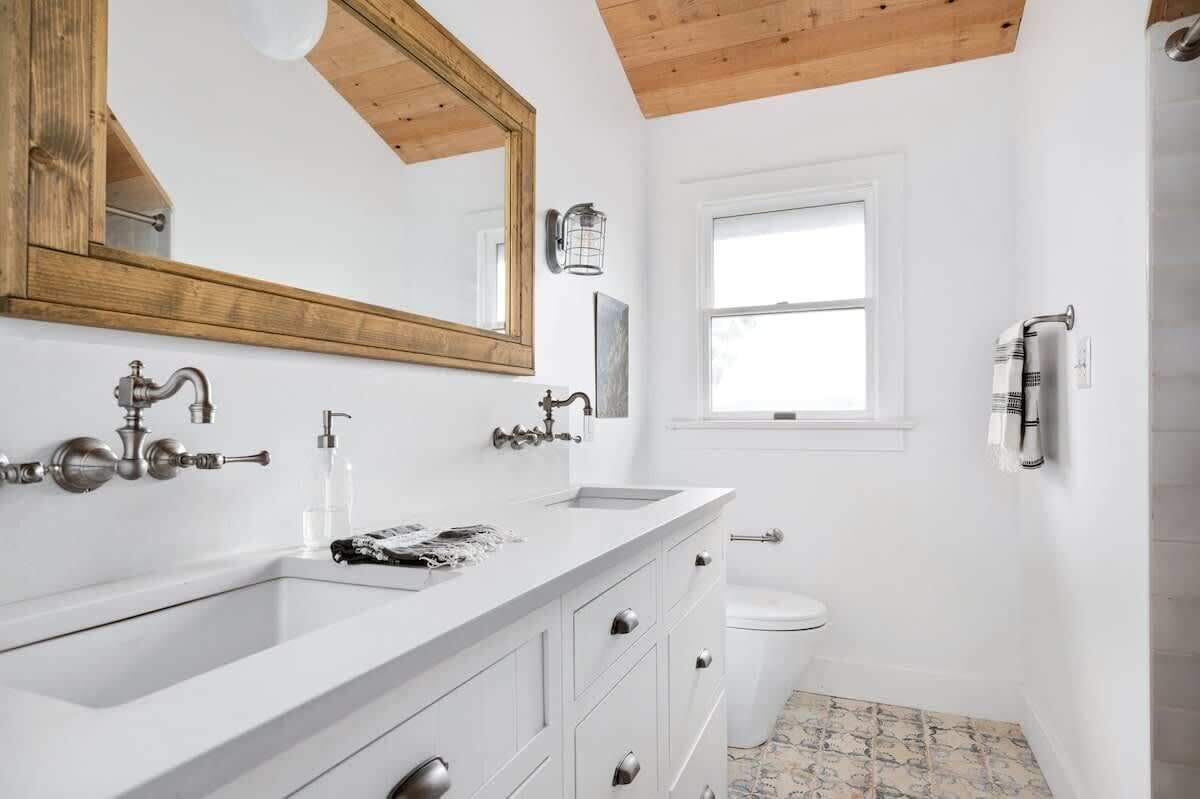 A bathroom with a white sink, double vanity, and a toilet. There is a window providing natural light and a wooden ceiling.