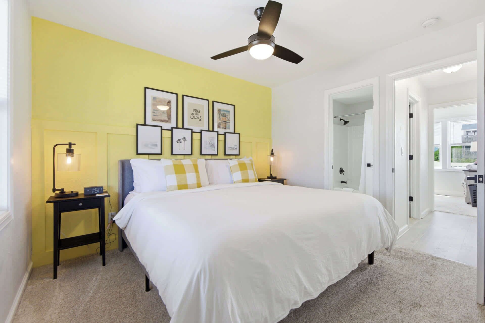 A bedroom with a bed, a nightstand, a wall clock, framed pictures, a ceiling fan, and a bathroom visible through an open door.