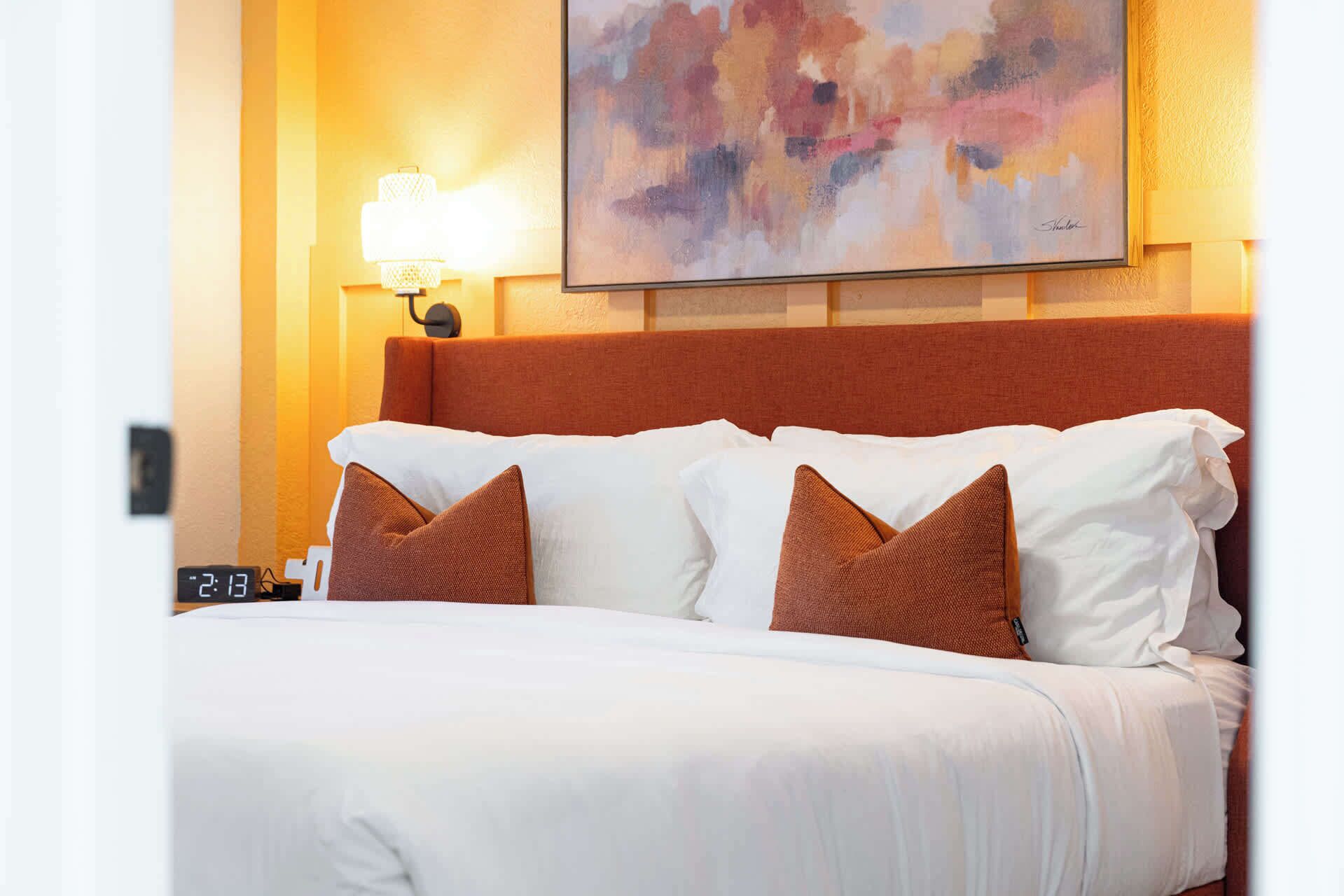 A hotel room with a bed, pillows, a wall clock, and a painting.