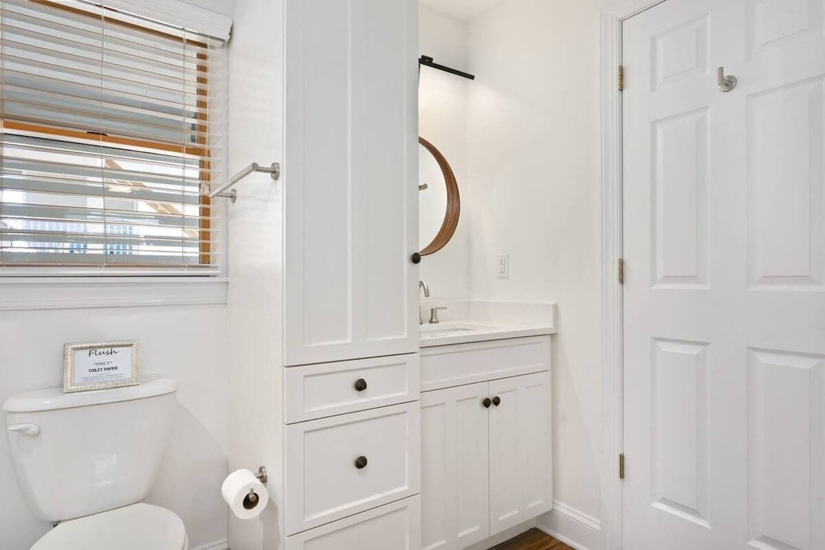 A bathroom with a toilet, a roll of toilet paper, a sign on the toilet, a sink, a mirror, and a door.