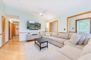 A spacious living room with a beige sofa, a coffee table, and a flat-screen TV mounted on the wall. - Pool, Hot Tub, Quick Walk to the Beach, EV Charger (Corolla)