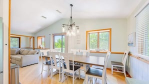 A spacious dining area with a long table and white chairs, a sofa, and a kitchen area with a refrigerator.