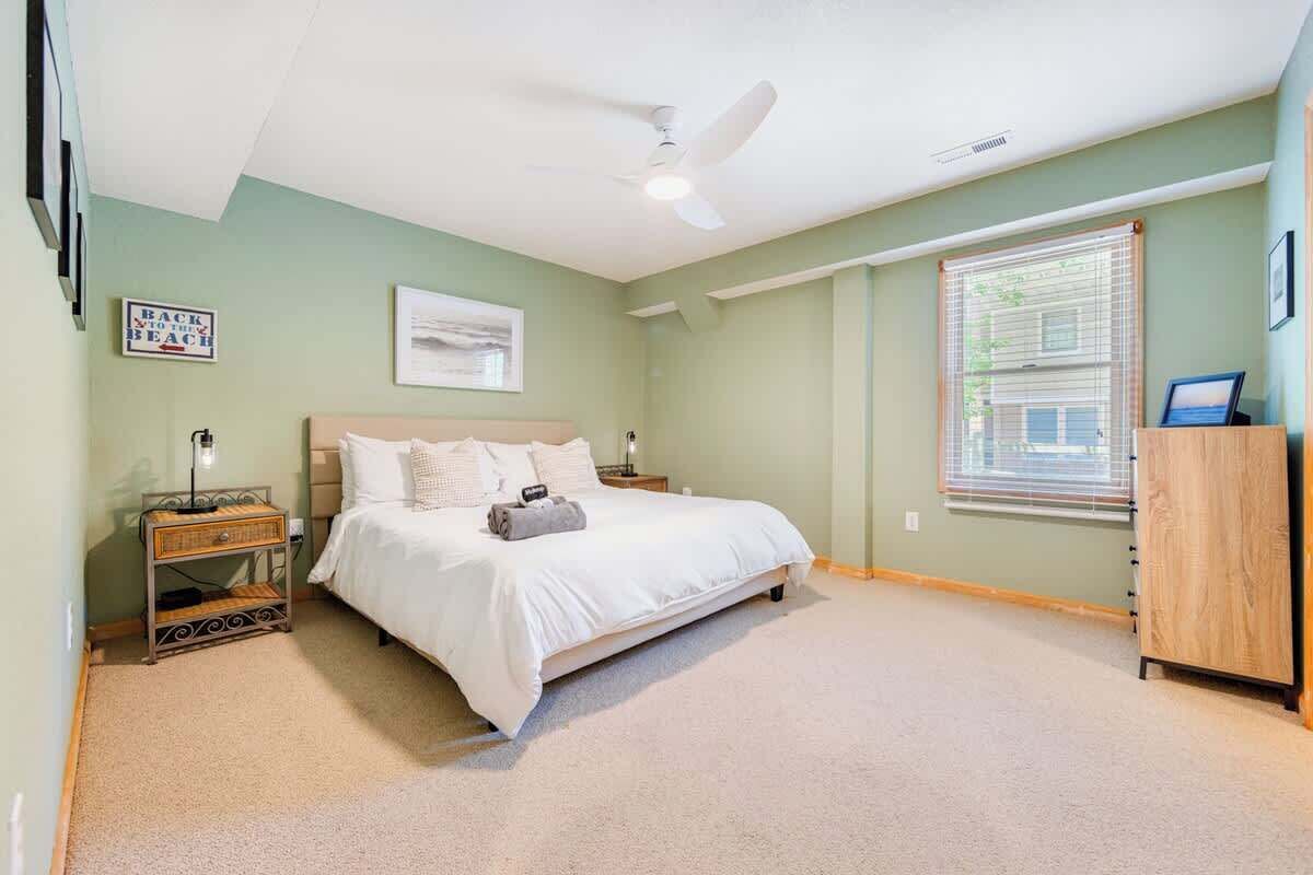 Een slaapkamer met een bed, nachtkastje, dressoir, plafondventilator en een raam met jaloezieën.