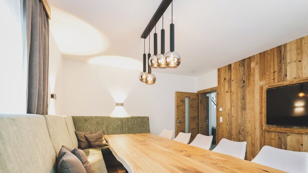 A modern dining area with a wooden table and white chairs, a sofa with cushions, and a wooden accent wall with a TV. - Chalet Carina by we rent (Zell am See)