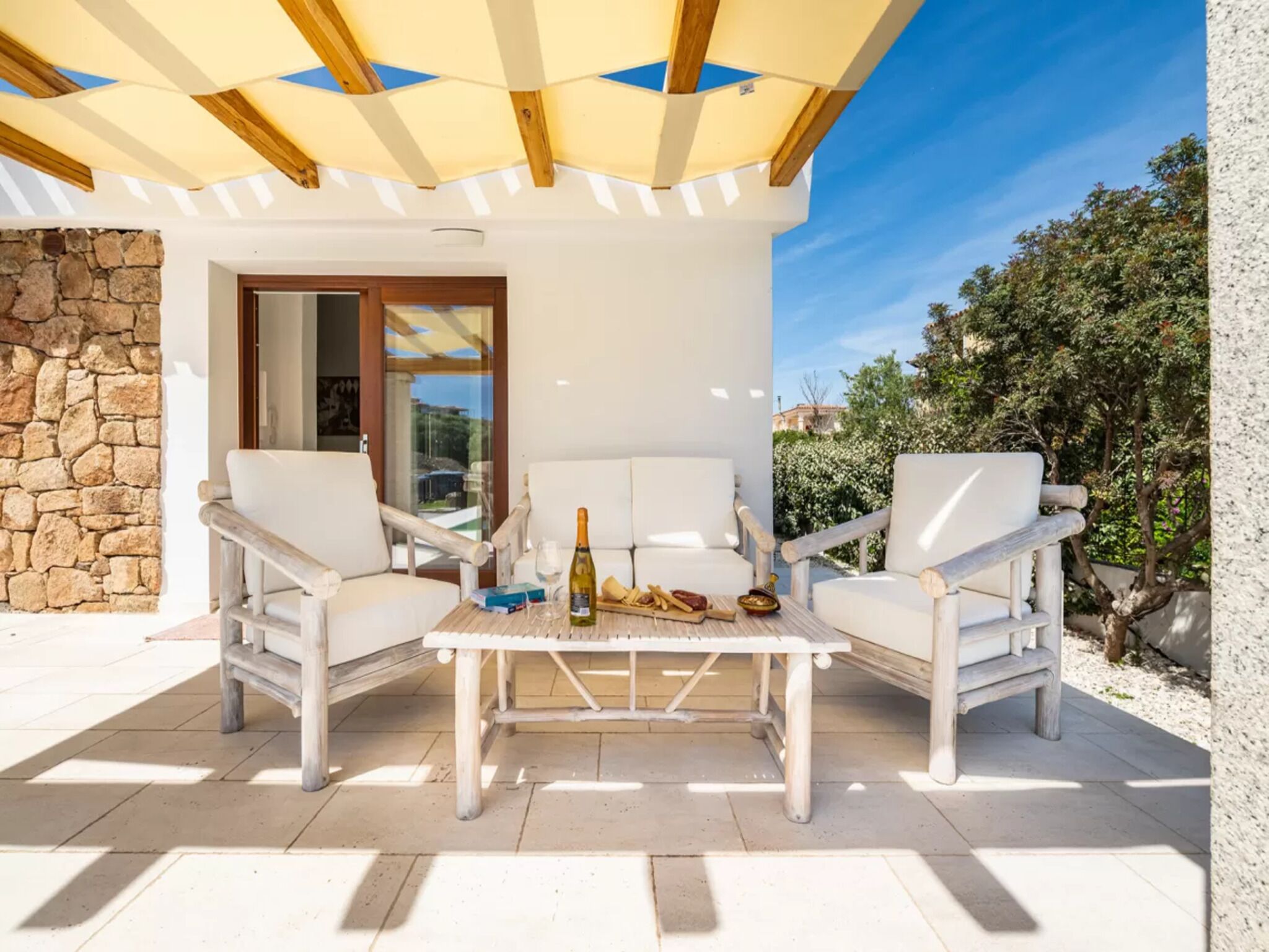 A patio with white furniture, a table with a bottle and glasses, and a stone wall.