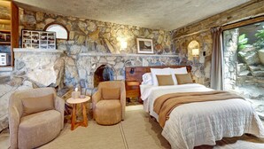 A bedroom with stone walls, a bed, a chair, a small table, and a fireplace. - Casa Grotto, 1 Bed Stone Cave Cottage, Panoramic Ocean View, Stone Shower, & Gym (San Marcos)