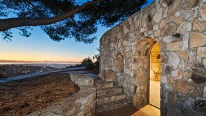 A stone building with an arched doorway, steps leading up to it, and a view of a distant cityscape at dusk. - Casa Grotto, 1 Bed Stone Cave Cottage, Panoramic Ocean View, Stone Shower, & Gym (San Marcos)