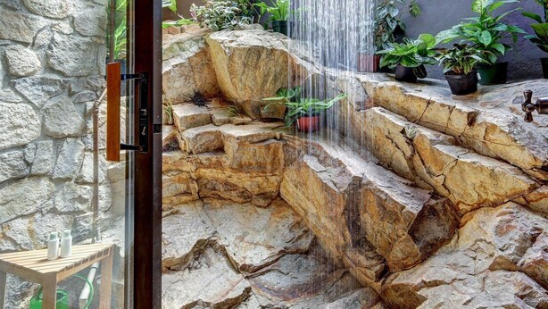 A stone-walled bathroom with a waterfall shower, potted plants, and a wooden bench. - Casa Grotto, 1 Bed Stone Cave Cottage, Panoramic Ocean View, Stone Shower, & Gym (San Marcos)