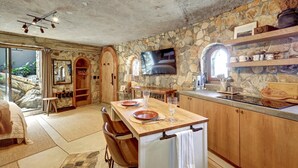 A rustic kitchen with stone walls, wooden cabinets, and a large TV. - Casa Grotto, 1 Bed Stone Cave Cottage, Panoramic Ocean View, Stone Shower, & Gym (San Marcos)