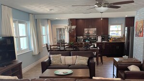 A spacious living room with a brown leather sofa, a dining area with wooden chairs, a kitchen with dark cabinets, and a ceiling fan.