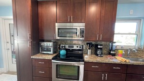 A modern kitchen with dark wooden cabinets, stainless steel appliances including a microwave, toaster oven, kettle, and coffee maker, granite countertops, and a sink with a faucet.
