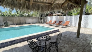 A pool area with a thatched roof, lounge chairs, and a table with chairs.