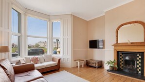 A living room with a fireplace, a sofa, a television, and large windows. - Heaton House Apartment - North East Escapes (Tynemouth)
