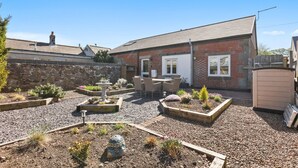 A courtyard with a stone wall, a brick building, a wooden fence, and a garden with various plants and a decorative fountain.