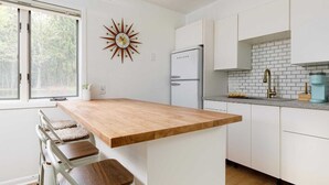 A modern kitchen with a wooden island, white cabinetry, a sunburst wall clock, and a view of trees through the window.