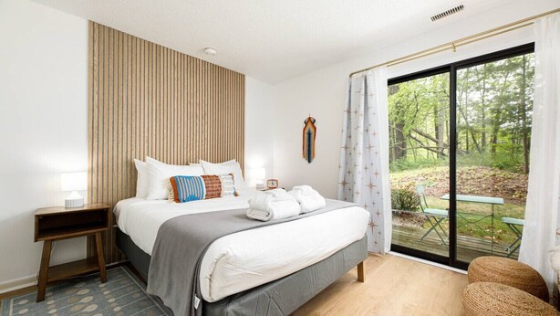 A modern bedroom with a large bed, a bedside table, a nightstand, a window with a view of the outdoors, and a wooden floor.