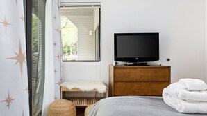 A bedroom with a bed, a TV on a wooden dresser, a window with curtains, a small bench, and a basket.