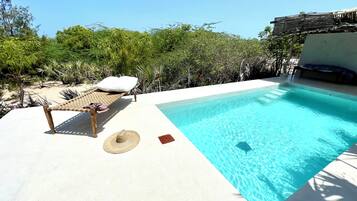 Una piscina con una silla de madera y un sombrero de paja, rodeada de follaje.