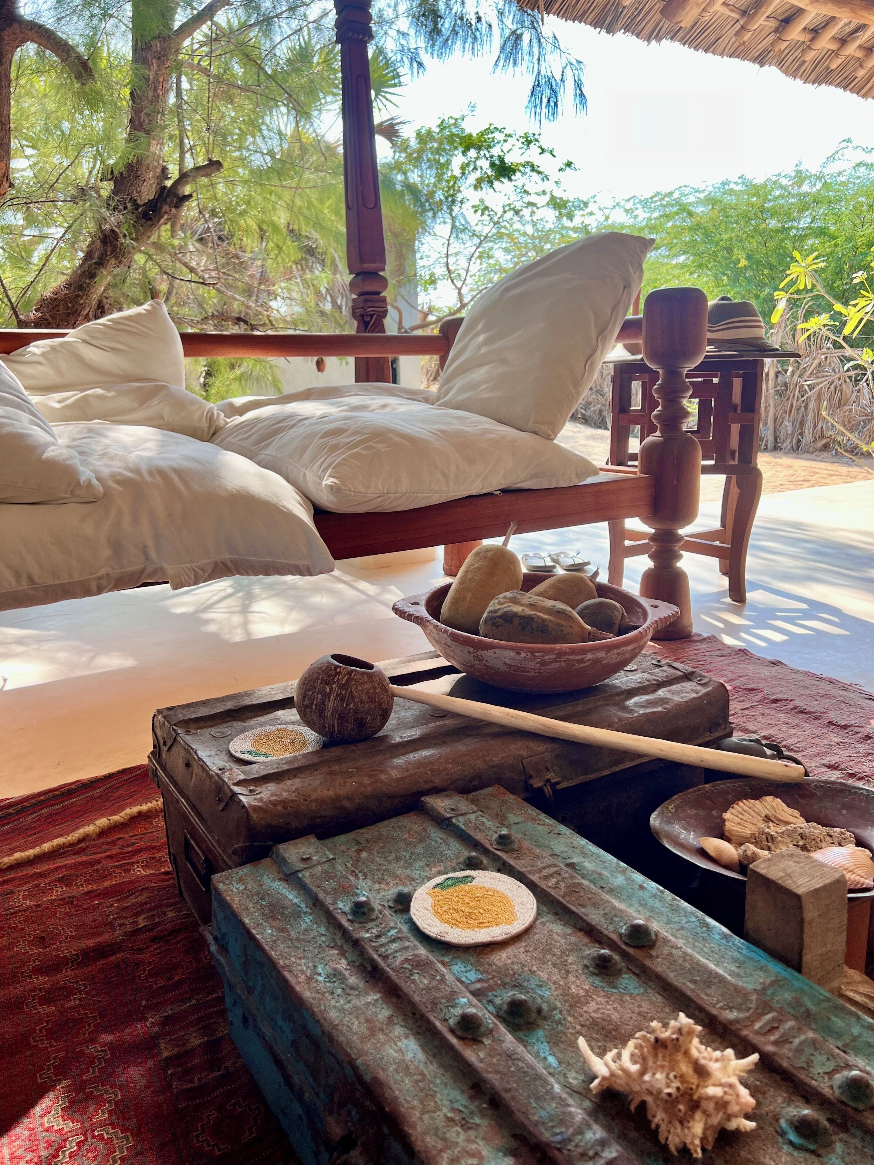 Un lit avec des oreillers blancs et une tête de lit en bois, une table rustique sur laquelle se trouve un saladier rempli de fruits et une cuillère en bois, et un tapis rouge à motifs.