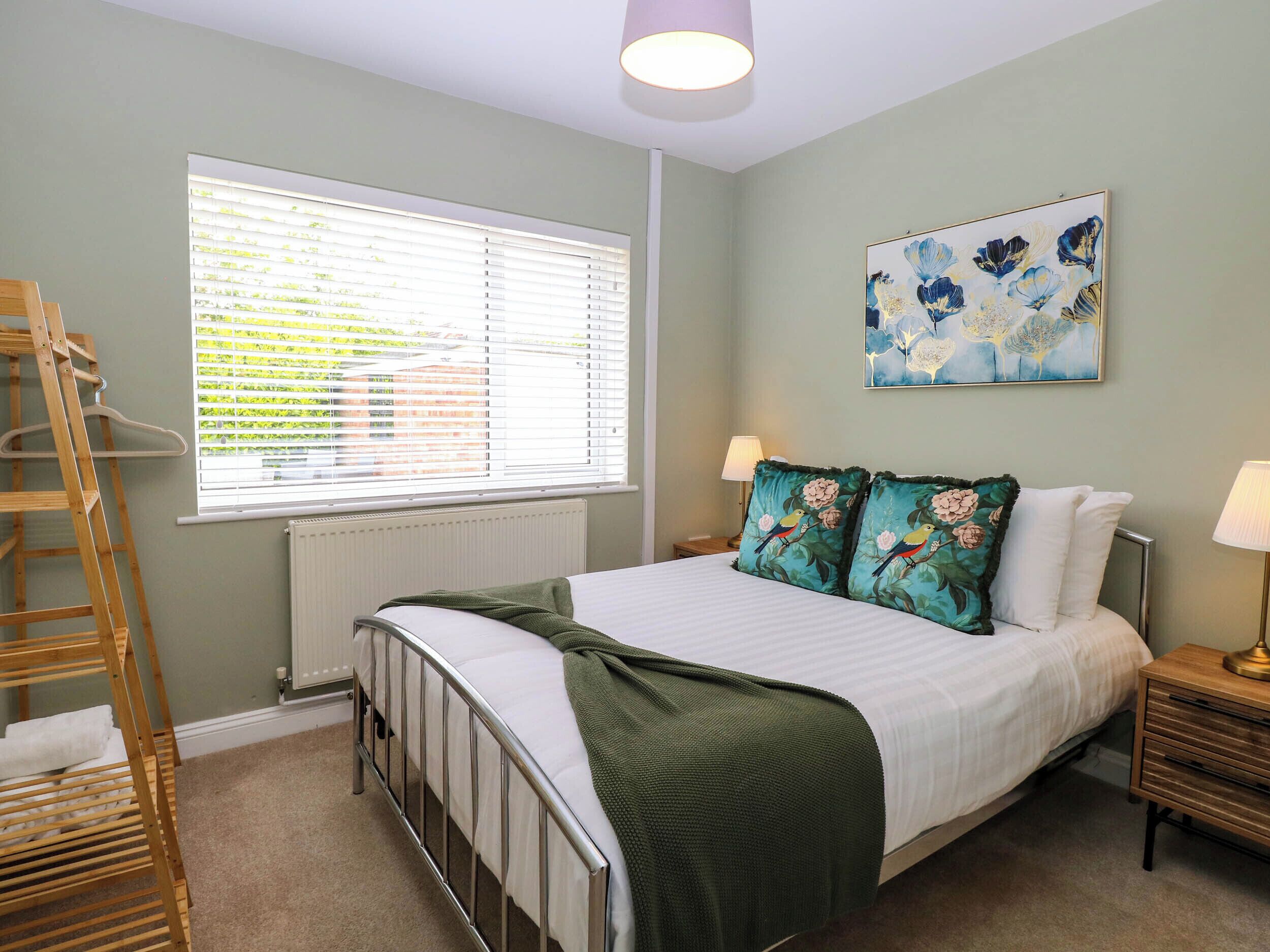 A bedroom with a bed, a radiator, a window with blinds, a painting, a nightstand, and a wooden ladder.