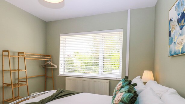 A hotel room with a bed, a window with blinds, a radiator, a wooden shelf unit, and a painting on the wall. - Little Lodge (King's Lynn)