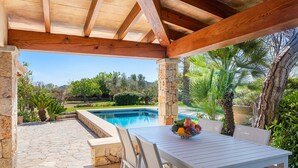 Un patio cubierto con una piscina de piedra, mobiliario blanco y vistas a un jardín paisajístico.
