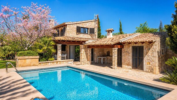 Una villa de piedra con piscina, mobiliario exterior y un jardín con árboles en flor.