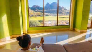 Una habitación con un ventanal grande que ofrece vistas a la montaña, un sofá con cojines y una persona sentada en el suelo.