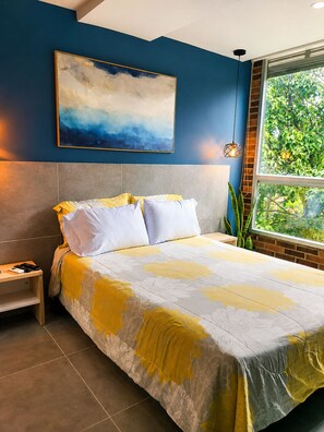 A bedroom with a bed, a nightstand, a painting, and a window with a view of greenery. - Stylish Laureles Loft in Medellin (Medellín)