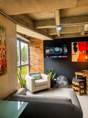 Uma sala de estar moderna com um sofá branco, uma mesa de centro de vidro, parede de tijolos, uma janela grande e uma TV fixada exibindo diversos canais.
