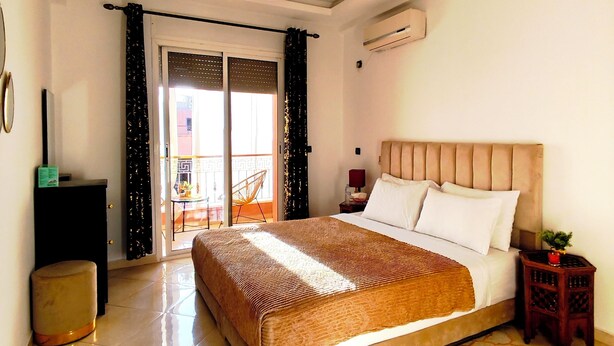 A bedroom with a large bed, a bedside table, a nightstand, a lamp, a small stool, a decorative rug, a ceiling light fixture, and a sliding glass door leading to a balcony.