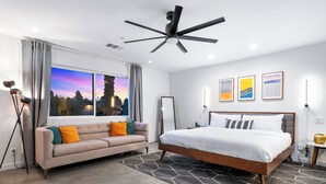 A modern bedroom with a bed, a sofa, a large window with a view, and a ceiling fan.