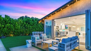 An outdoor patio with modern furniture, a kitchen area visible through glass doors, and a well-maintained lawn.