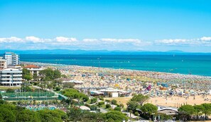 A beach with numerous umbrellas and people, a clear blue sky, and buildings in the background.